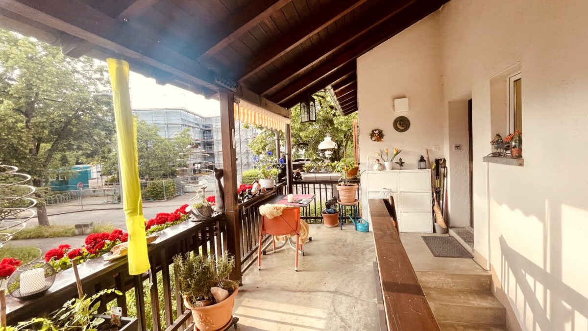 Ein gemütlicher Balkon mit Topfpflanzen, roten Blumen am Geländer, einem kleinen Tisch und Stühlen und Sonnenlicht, das unter einem Holzdach hindurchscheint.