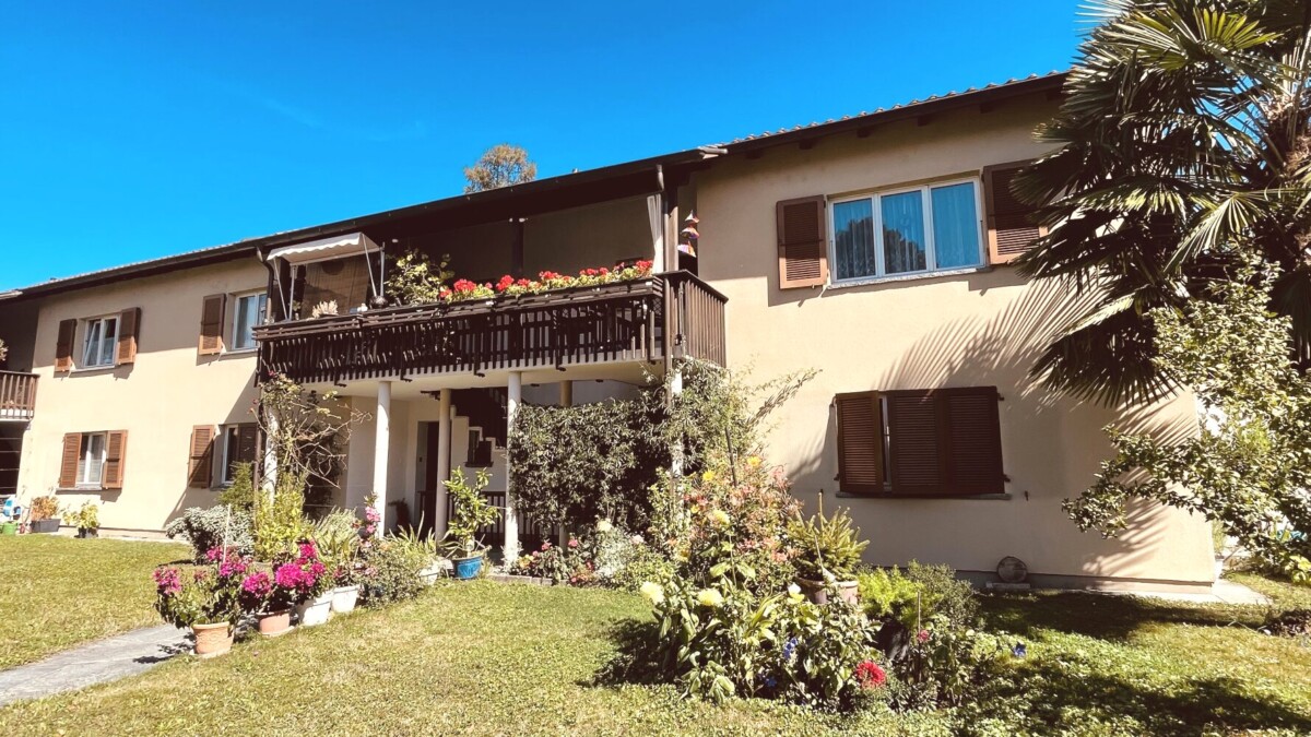 Ein zweistöckiges beigefarbenes Haus mit hölzernen Fensterläden und einem blumengeschmückten Balkon, umgeben von einem üppigen Garten und Topfpflanzen unter einem strahlend blauen Himmel.