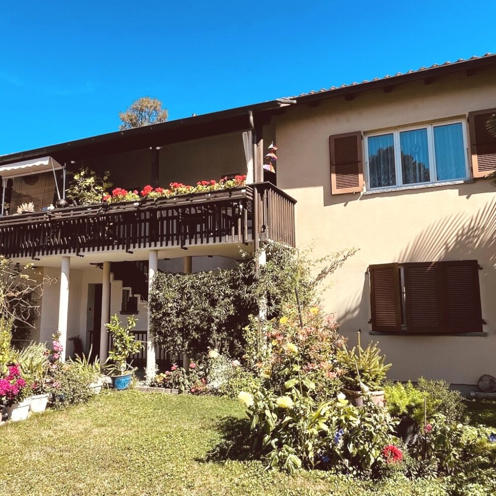 Ein zweistöckiges beigefarbenes Haus mit hölzernen Fensterläden und einem blumengeschmückten Balkon, umgeben von einem üppigen Garten und Topfpflanzen unter einem strahlend blauen Himmel.