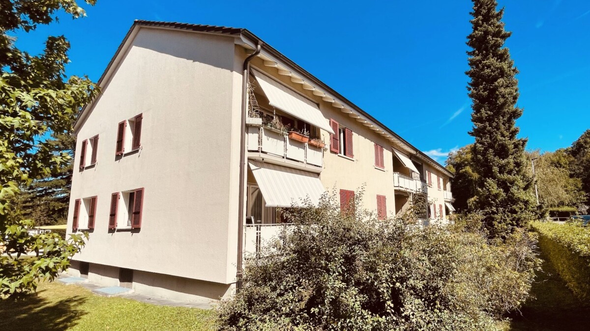 Ein zweistöckiges beigefarbenes Wohnhaus mit roten Fensterläden und gestreiften Markisen, umgeben von Grün unter einem klaren blauen Himmel.