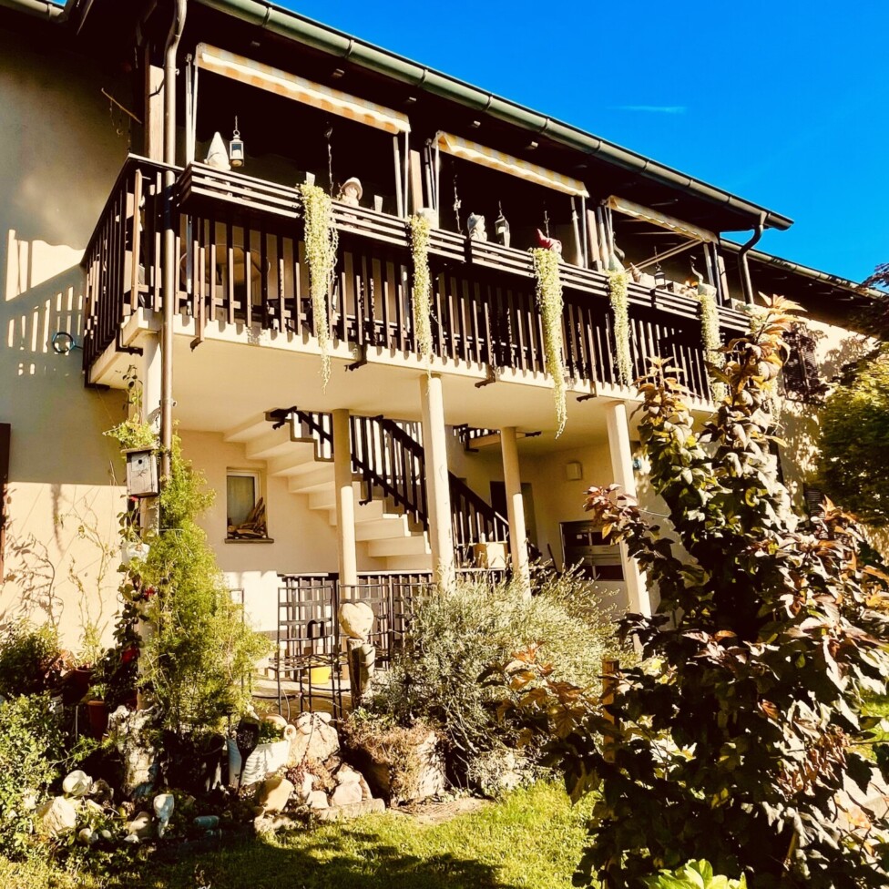 Ein zweistöckiges cremefarbenes Haus mit braunen Fensterläden, einem dunklen Holzbalkon und einer Treppe, umgeben von grünem Gras, Bäumen und Pflanzen, unter einem strahlend blauen Himmel.