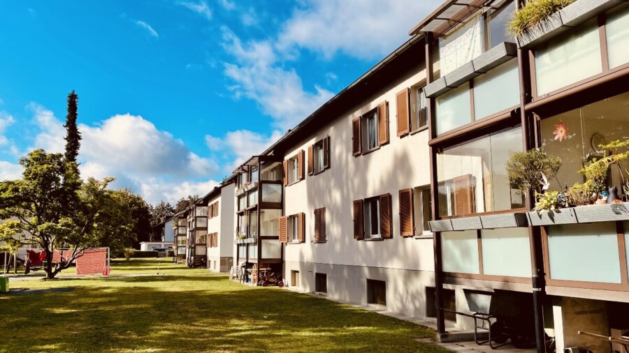 Eine Reihe moderner Wohnhäuser mit Balkonen, die auf einen grasbewachsenen Innenhof, Bäume und einen strahlend blauen Himmel mit vereinzelten Wolken blicken.