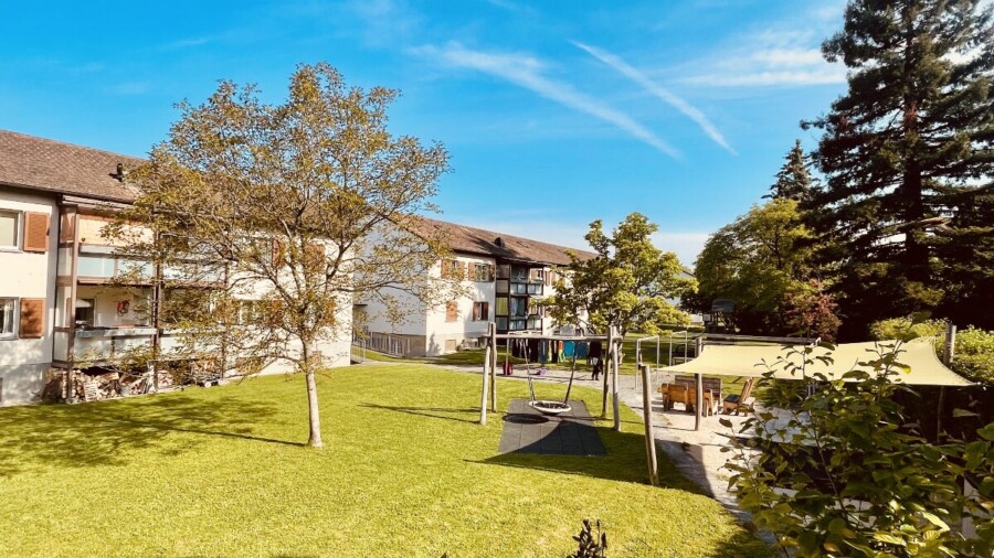 Ein sonniger Innenhof mit grünem Gras, Bäumen, einem Spielplatz mit Schaukeln und einem gelben Schattendach neben einem weißen Wohngebäude mit Balkonen. Blauer Himmel mit ein paar Schäfchenwolken darüber.