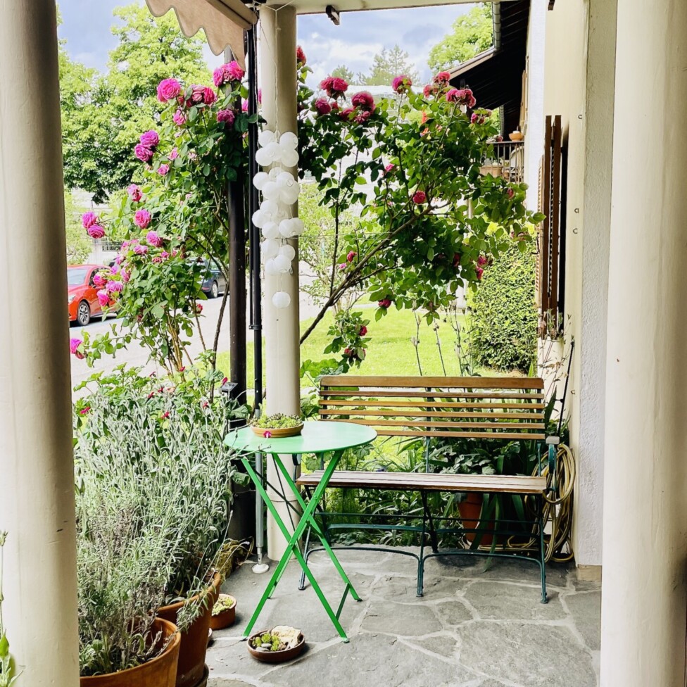 Eine gemütliche Veranda mit Topfpflanzen, einem grünen Klapptisch und -stuhl sowie einer Holzbank. An den Sträuchern blühen rosa Blumen, und im Hintergrund parkt ein rotes Auto auf der Straße. Steinfliesen bedecken den Boden.